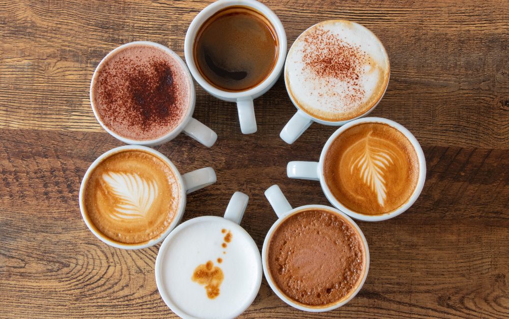 Different variants of coffee served in white mugs sitting on a wooden table.